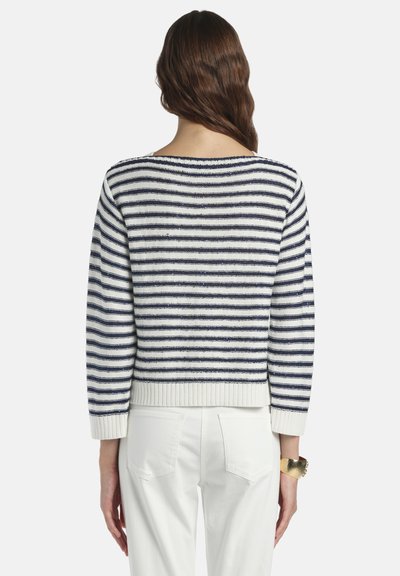 Femme aux longs cheveux bruns portant un pull à rayures horizontales bleu marine et blanc et un pantalon blanc, debout les bras détendus le long du corps.