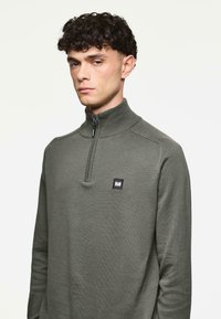 Mies, jolla on kihara tummat hiukset, pukeutuneena pitkähihara oliivinvihreään neljännesvetoketjupaitaan, jossa on pieni suorakulmainen logo rinnassa.