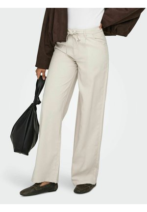 Person som har på seg beige vide bukser, brun jakke og mørke loafers, holder en svart knyttet skinnveske ved siden av seg.