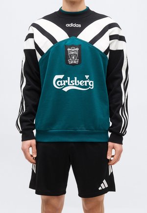 Personne portant un sweat-shirt vert, noir et blanc Adidas Liverpool Football Club avec le logo Carlsberg et un short noir Adidas.