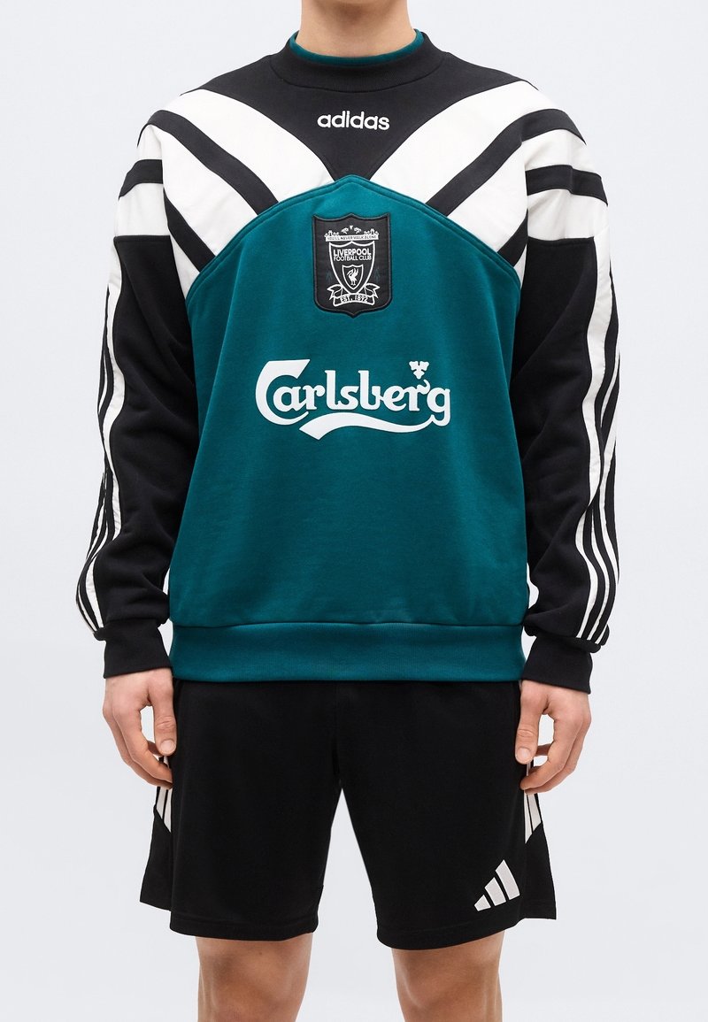 Personne portant un sweat-shirt vert, noir et blanc Adidas Liverpool Football Club avec le logo Carlsberg et un short noir Adidas.
