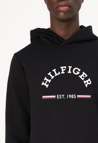 Sort hættebluse med et fronttryk, der viser "HILFIGER" i hvid bue-skrift og "EST. 1985" nedenunder, akkompagneret af røde og blå striber.