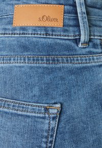 Jeans en denim bleu clair avec une surface texturée, présentant une étiquette en cuir marron embossée "s.Oliver" près de la taille et des détails de couture standard.
