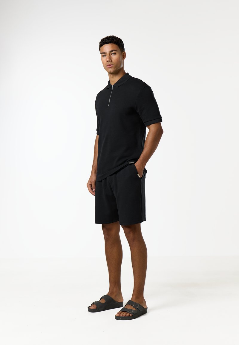 Homme debout portant un polo noir à manches courtes avec fermeture éclair, un short noir et des sandales noires sur fond blanc uni.