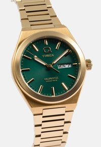 Zlatý quartzový hodinky Timex s textúrovaným zeleným ciferníkom, hodinovými značkami a zobrazením dňa. Ploché zlaté náramky dokončujú dizajn.