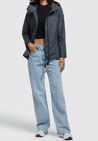Dunkelgraue, leichte Jacke mit Kapuze, Reißverschluss und Taschen, kombiniert mit hellblauen, weit geschnittenen Jeans und gemusterten Sneakern.