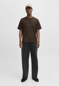 Brun kortærmet T-shirt med "JACK & JONES" logo, parret med løstsiddende sorte denimjeans og sorte sko, båret med en beige cap.