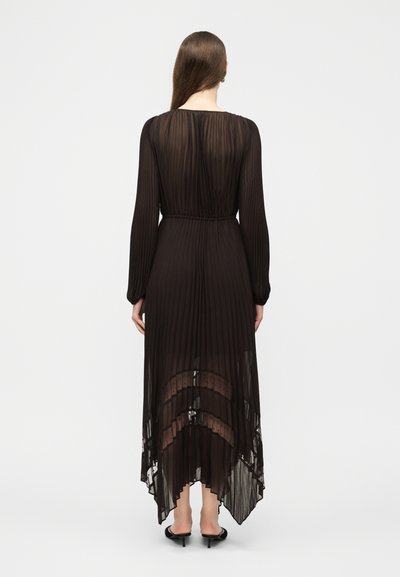 Γυναίκα που φοράει μακρύ, διαφανές, μαύρο πλισέ φόρεμα με μακριά μανίκια και ασύμμετρο τελείωμα, φαίνεται από πίσω με φόντο ένα απλό φόντο.