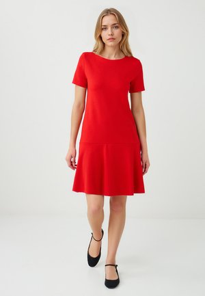 Modèle portant une robe rouge à manches courtes avec une jupe évasée, se tenant devant un fond blanc uni, regardant directement le spectateur.