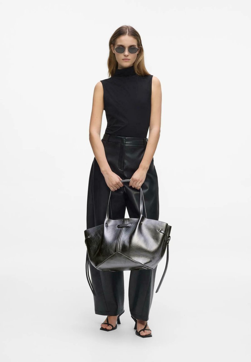 Femme portant un top noir sans manches, un pantalon en cuir taille haute, des sandales et des lunettes de soleil, tenant un grand sac à main en cuir noir à deux poignées.