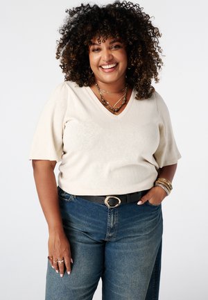 Femme souriante aux cheveux bouclés porte un t-shirt beige à col en V, un jean bleu, une ceinture noire, des colliers superposés, des bracelets et des bagues, main dans la poche.