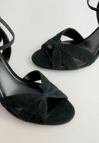 Sorte ruskinds højhælede sandaler med krydsede stropper foran og en glat sort indvendig. Åben tå-design med en slank ankelslyd.