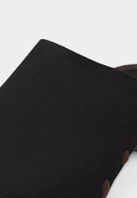 Chaussure noire sans lacets avec dessus lisse et clous métalliques ronds visibles le long du côté près de la semelle.