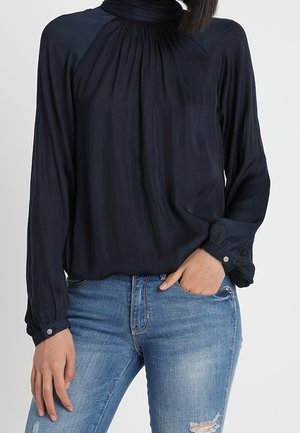 Femme portant un chemisier bleu marine à manches longues avec un col et des poignets froncés, associé à un jean bleu clair déchiré.