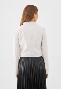 Ljusrosa läderkappa med kort modell och långa ärmar, figursydd design och platt krage, i kombination med en svart veckad midikjol.