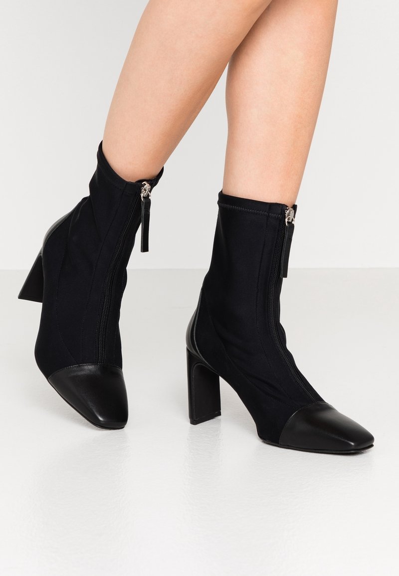 Bottines noires avec un dessus en tissu extensible, un bout en cuir verni et une fermeture éclair latérale avec un tirant décoratif. Design à talon bloc.