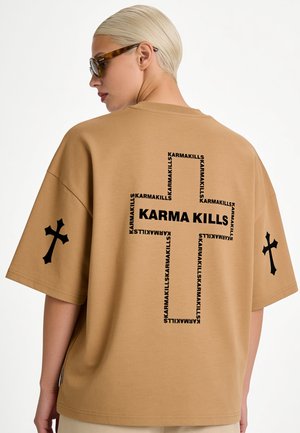 Personne aux cheveux blonds courts et lunettes de soleil portant un t-shirt oversized beige avec des motifs croix noirs et le texte "KARMA KILLS" au dos et aux manches.