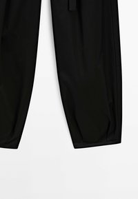 Partie inférieure d’un pantalon noir à jambes resserrées, présentée sur un fond blanc.