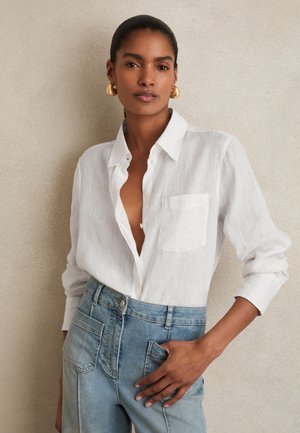 Donna con i capelli tirati indietro, indossa orecchini a cerchio dorati, camicia bianca con bottoni e jeans azzurri a vita alta, in piedi contro un muro semplice.
