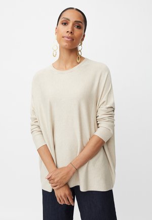Maglione oversize beige chiaro con maniche lunghe e polsi a coste, abbinato a pantaloni in denim scuri. Gli accessori includono grandi orecchini circolari dorati.