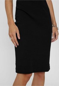 Robe noire côtelée de longueur genou en tissu tricoté, présentant une silhouette ajustée et une texture lisse, avec une main reposant sur le côté.