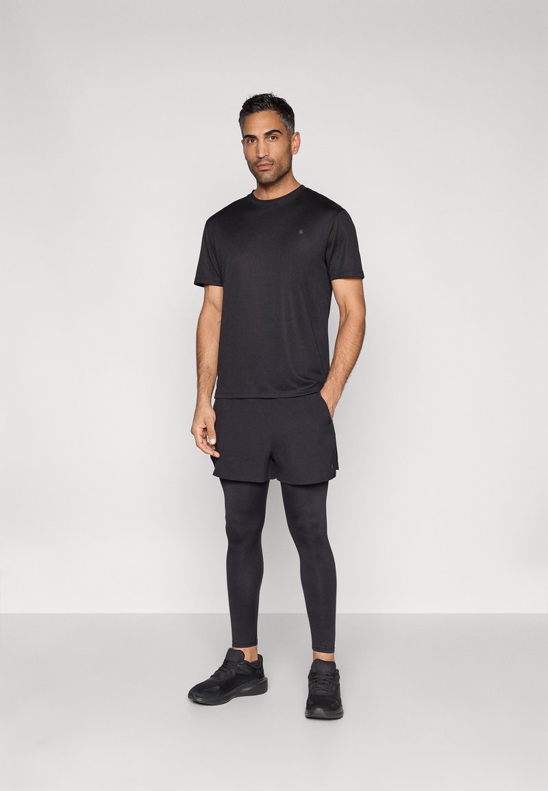 Férfi áll, fekete sportpólóban, rövidnadrágban a leggings fölött, és fekete tornacipőben, egyszerű világos háttér előtt.