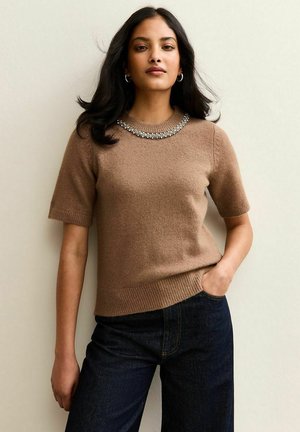 Suéter corto marrón de manga corta con una textura suave, que presenta un escote adornado con joyas. Combinado con jeans de color azul oscuro.