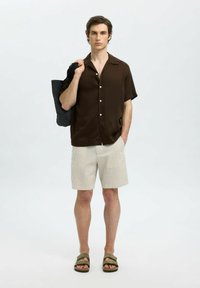 Brun kortärmad skjorta med knappar i kombination med beiga randiga shorts. Modellen håller en mörk axelväska och har på sig olivgröna sandaler.