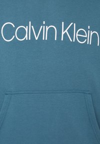 Miętowa bluza z kieszenią z przodu, z dużym, białym logo tekstowym "Calvin Klein" na piersi. Gładka faktura materiału.