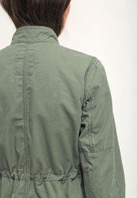 Khurty zielony lekki kurtka z podniesionym kołnierzem, zebranym w talii i gładkim materiałem, z subtelnymi detalami sticznymi i bez widocznych elementów metalowych.