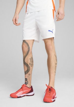 Shorts de sport blancs avec des accents orange et un logo Puma bleu. Baskets rouge vif avec des détails blancs et marron. Tatouages détaillés sur les jambes visibles.