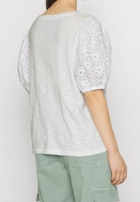 Bílý top s texturovanými nabíranými rukávy s vyšívanými detaily. Zadní strana je jednoduchá a o něco delší, spárovaný s zelenými cargo kalhotami.