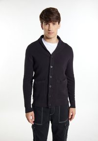 Cardigan scuro e strutturato con colletto a scialle, sette bottoni sul davanti e due tasche frontali. Indossato sopra una camicia bianca.