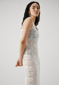 Robe au crochet blanche présentant un motif géométrique, une silhouette ajustée et des bretelles spaghetti, avec une texture ajourée complexe tout au long.