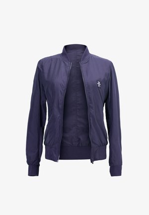 Junge Frau mit langen dunklen Haaren, die eine marineblaue Jacke über einem grauen Hoodie und blauen Jeans trägt, steht mit den Händen in den Jackentaschen und schaut seitlich.
