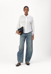 Chemise blanche à manches longues et col, portée avec un jean bleu délavé à jambes larges et un sac à main en osier noir ; ballerines noires.