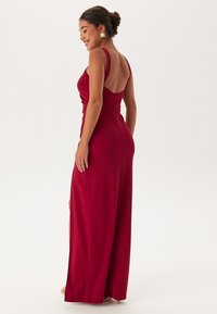Robe longue et ajustée en rouge profond, dotée d'un dos plongeant, de fines bretelles et d'une texture lisse. Une fente sur le côté ajoute un détail au design.