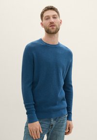 Suéter azul de punto con escote redondo y mangas largas, que presenta un patrón texturizado, combinado con jeans de color azul claro.