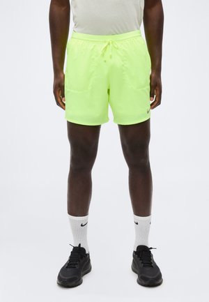 Personne portant un short de sport jaune vif avec cordon, des chaussettes blanches Nike montantes et des chaussures de sport noires, debout devant un fond neutre.