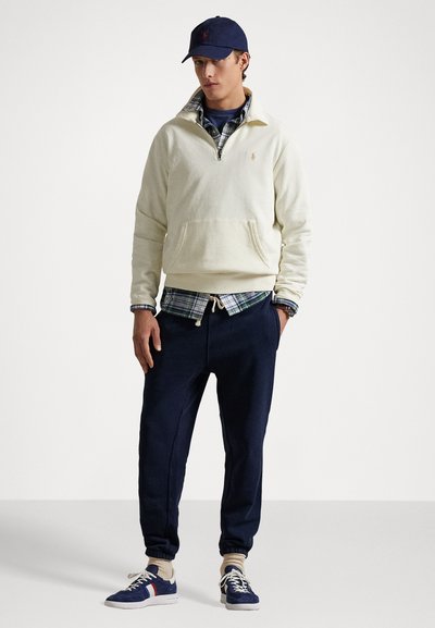 Lys kremfarget genser med halv glidelås, marineblå bukser med elastiske mansjetter, marineblå caps, og blå sneakers med røde og hvite striper. Rutete skjorte under.