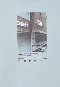 Camiseta de color azul claro con un estampado gráfico de una escena urbana texturizada, con el texto "Momentos de Calle Urbana" y los controles de reproducción debajo.