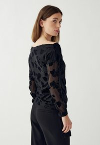 Fekete felső áttetsző, hosszú ujjakkal, virágmintás textúrával, kerek nyakkivágással és figura-követő szabással. Fekete nadrággal viselve.