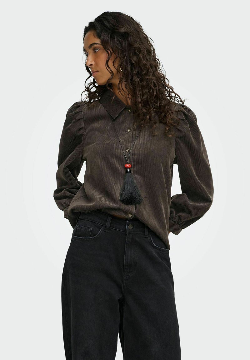 Jeune femme aux cheveux bouclés portant un chemisier marron foncé boutonné avec des manches bouffantes, un pantalon noir et un long collier à pompon noir avec une perle rouge.