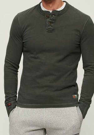 Man met donkergroen longsleeve henley-shirt met knoopsluiting en lichtgrijze joggingbroek, handen in de zakken, tegen een neutrale achtergrond.