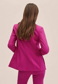 Kobieta o długich brązowych włosach, ubrana w dopasowany, jaskraworóżowy garnitur, stoi w pozycji z rękami na biodrach, zwrócona tyłem na neutralnym tle.