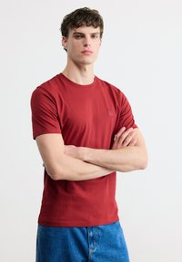 Νεαρός άνδρας με κατσαρά μαλλιά, που φοράει κόκκινο κοντομάνικο πουκάμισο και μπλε τζιν, στέκεται με σταυρωμένα χέρια μπροστά σε ένα απλό λευκό φόντο.