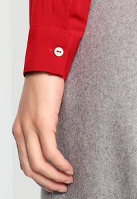 Poignet de chemise en coton rouge avec bouton blanc, main reposant sur un tissu gris texturé. Détails nets sur le tissu et les caractéristiques du bouton visibles.