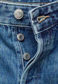 Gros plan sur un jean en denim bleu avec trois boutons métalliques argentés et une boutonnière ouverte sur une ceinture pliée.