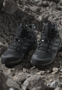 Bottines de randonnée montantes noires, avec une tige synthétique texturée, un embout protecteur et trois rayures argentées sur les côtés.