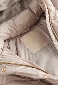 Ljust beige puffjacka med en slät innerväv, quiltat mönster och en gyllene etikett som står "NEXT THE FUTURE IS NOW EST. 1982." Gyllene dragkedja.